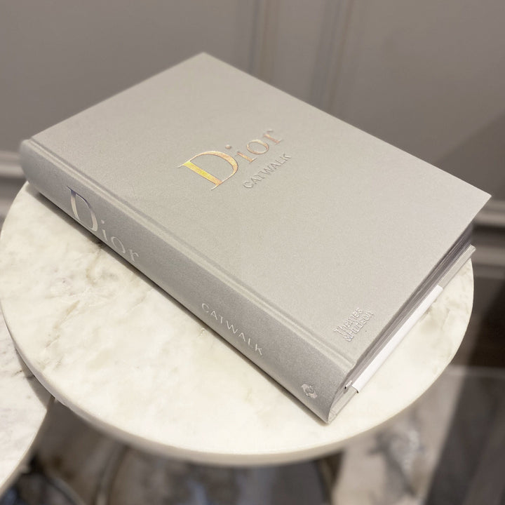 The Dior Catwalk Book in grey featuring silver metallic typography on a marble table. The perfect Dior Coffee Table Book.